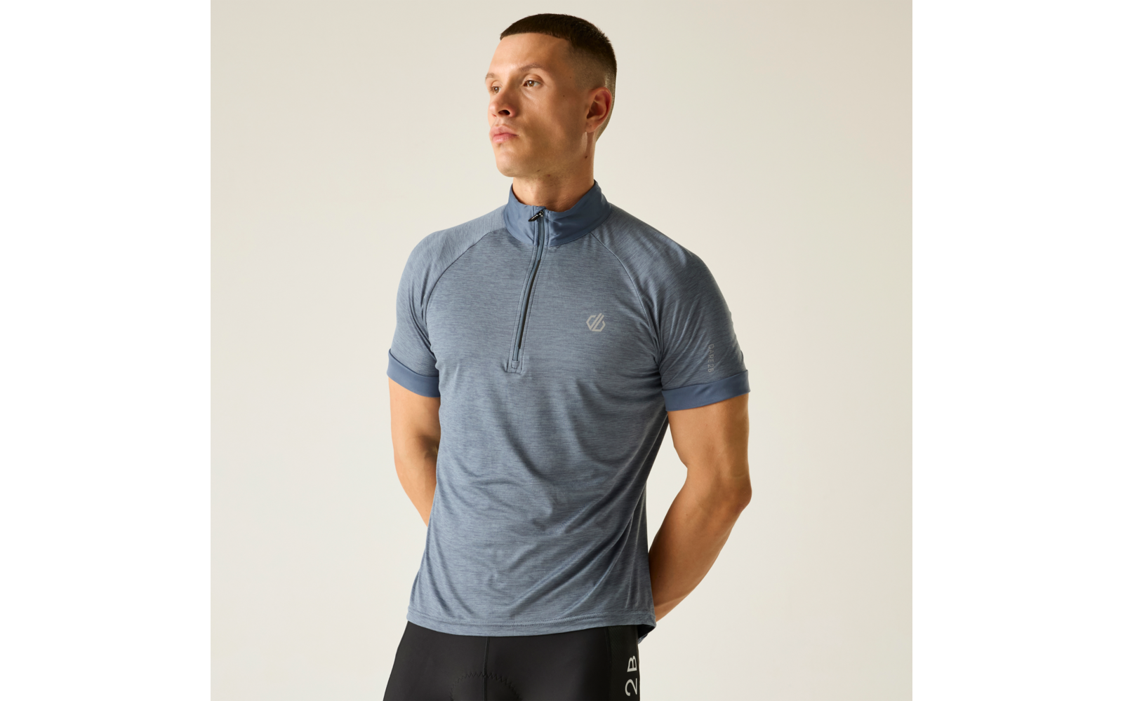 Person in blau-grauem, kurzärmeligem Sportshirt mit kurzem Reißverschluss und schwarzen Radshorts, Hände hinter dem Rücken, nach links schauend, heller einfarbiger Hintergrund