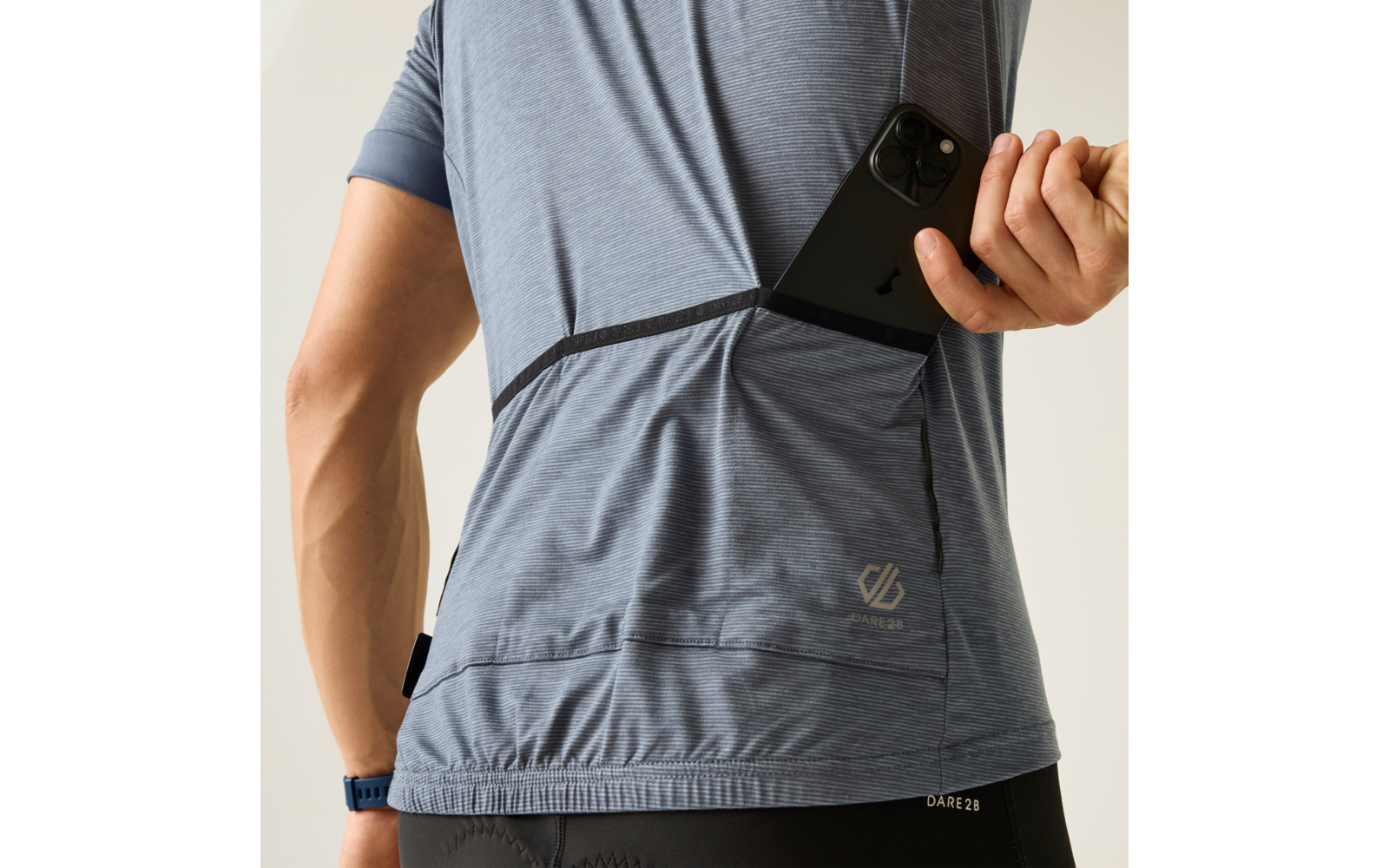 Rückenansicht einer Person in blaugrauem Sportshirt, die ein schwarzes Smartphone in eine Rückentasche steckt; sichtbarer Arm mit Armbanduhr, Logo auf dem Shirt und schwarze Sporthose