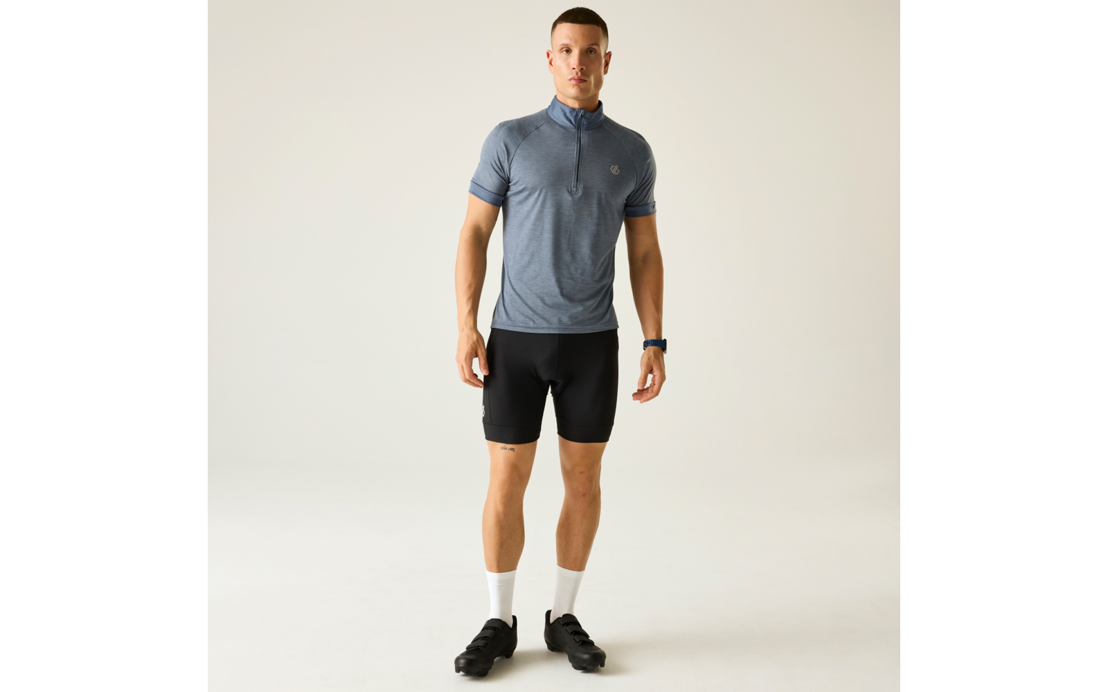 Mann in blauem, kurzärmeligem Fahrradtrikot mit Reißverschluss, schwarzer Radhose, weißen Socken und schwarzen Fahrradschuhen, steht frontal vor hellem Hintergrund