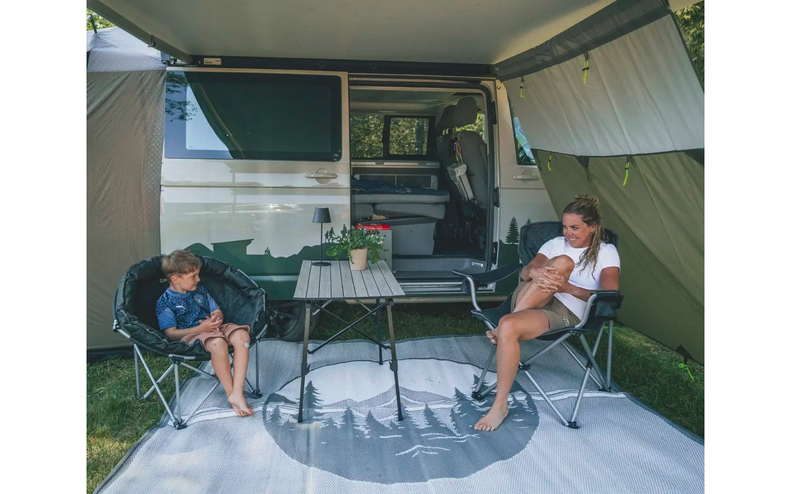 Zwei Männer sitzen vor einem Camper-Van mit geöffnetem Vorzelt, ein Campingtisch steht zwischen ihnen