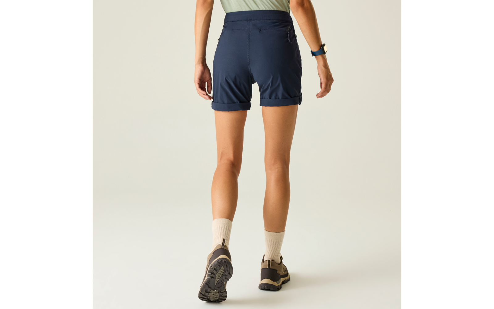 Rückansicht einer Person in dunkelblauen Shorts, hellgrünem Oberteil, hellen Socken und braunen Schuhen mit grober Sohle; Armbanduhr am linken Handgelenk