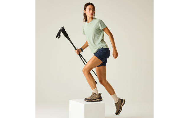 Person trägt grünes T-Shirt, blaue Shorts und Wanderschuhe, steht mit einem Fuß auf einem weißen Würfel und hält zwei Wanderstöcke, heller Hintergrund