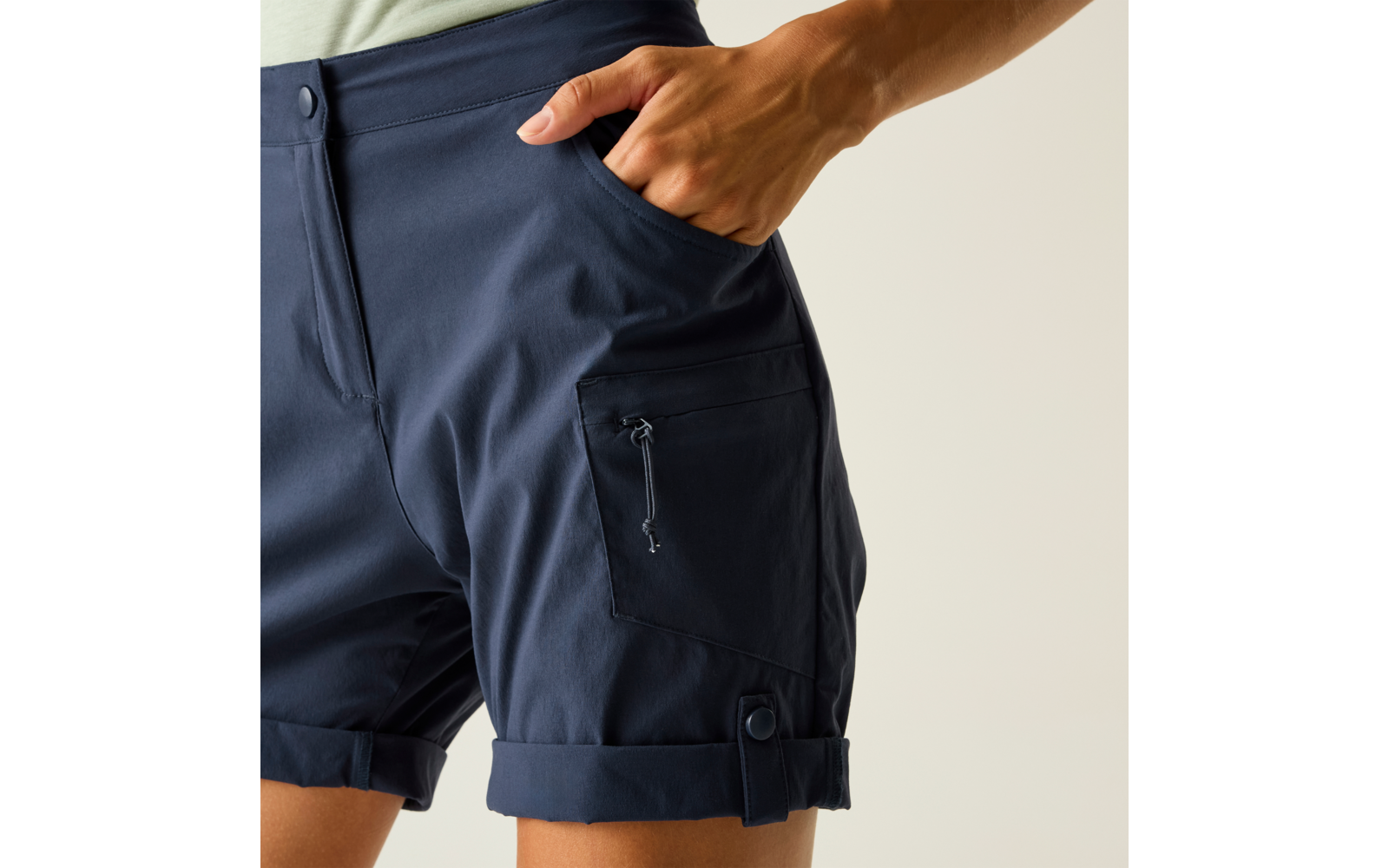 Nahaufnahme der Hüfte und oberen Oberschenkel einer Person in dunkelblauen Shorts; Hand in vorderer Tasche; seitliche Reißverschlusstasche und umgeschlagener Saum sichtbar