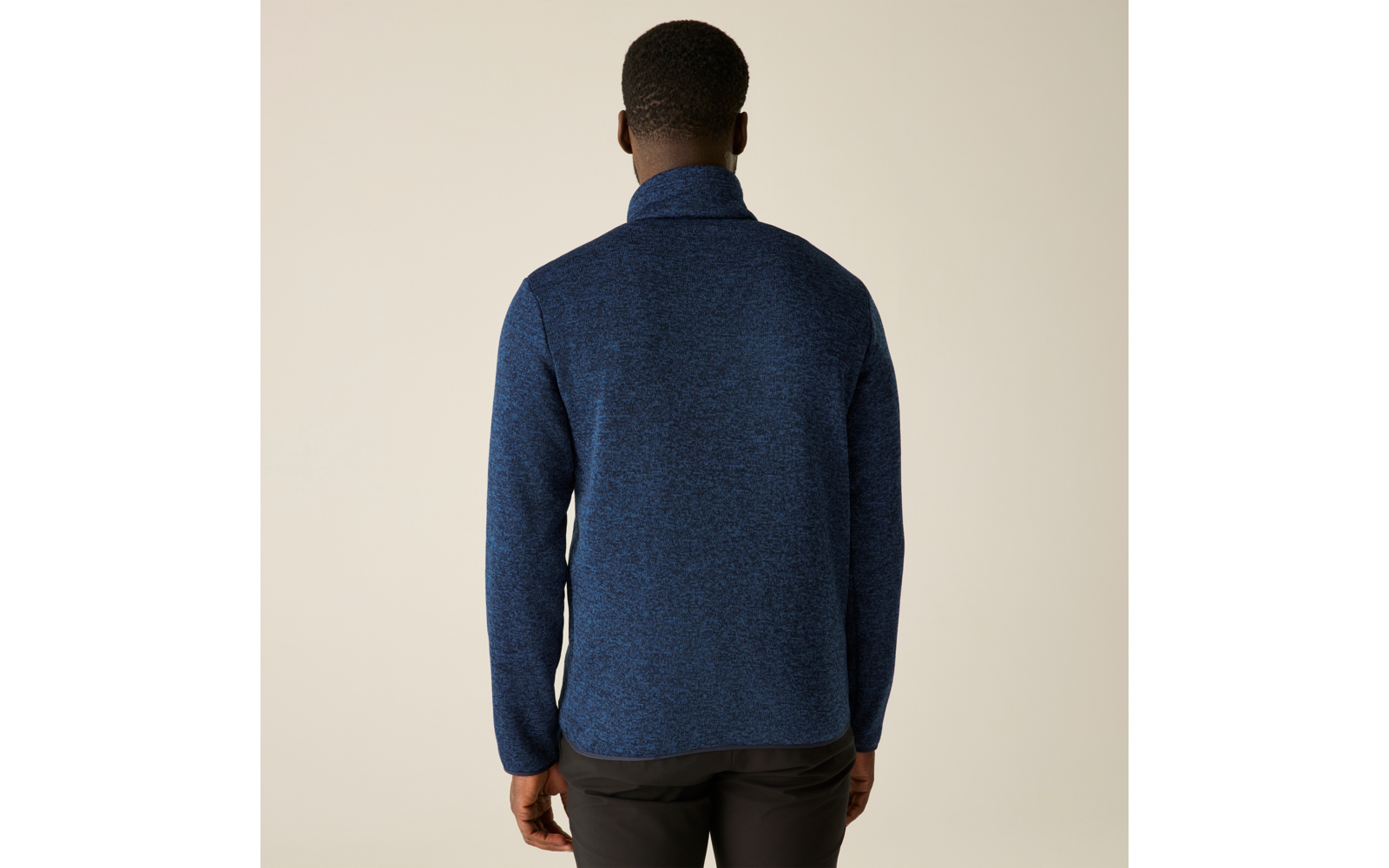 Persoon van achteren, draagt een donkerblauwe trui met opstaande kraag en een donkere broek voor een egaal lichte achtergrond