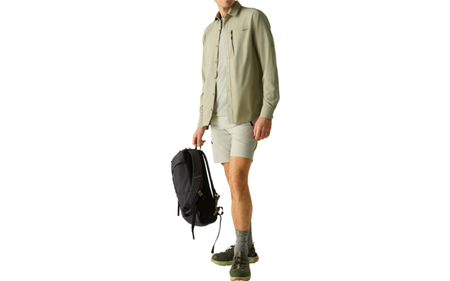 Stehender Mann in hellgrünem Hemd, hellen Shorts und Wanderschuhen, trägt schwarzen Rucksack in der rechten Hand, heller Hintergrund