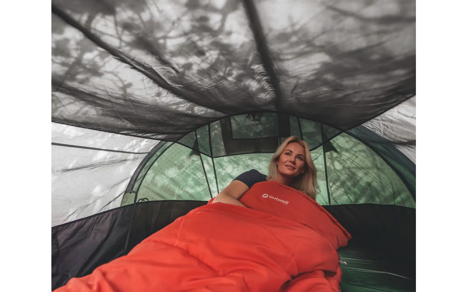 Frau legt sich lächelnd in ein Zelt, grüne Wiese im Hintergrund