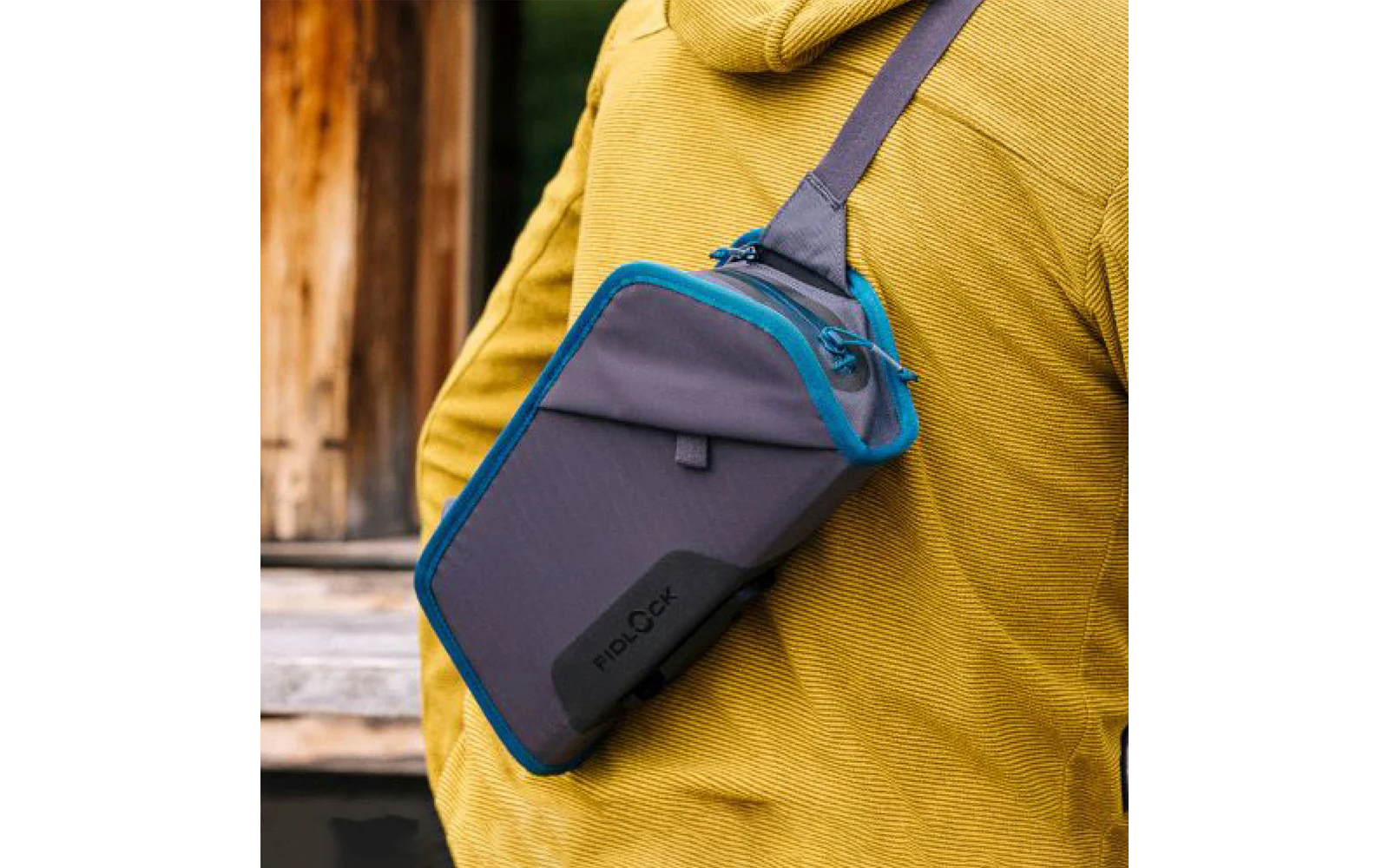 Person von hinten in gelber Jacke trägt eine graue Umhängetasche mit blauem Rand quer über dem Rücken; Holzwand und Pflanzen im Hintergrund