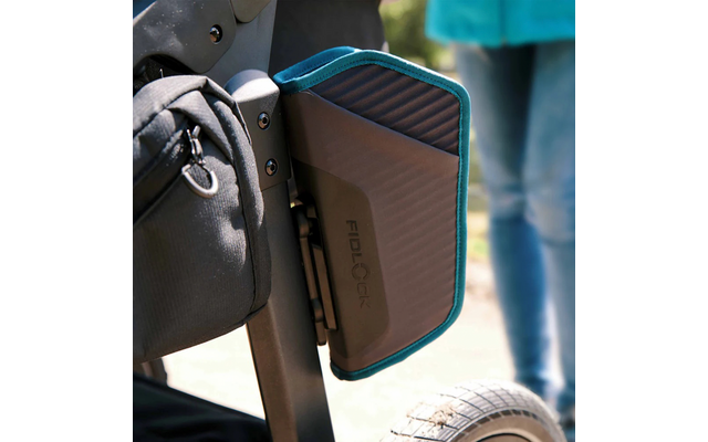 Nahe Aufnahme eines schwarzen, rechteckigen Fidlock‑Cases mit türkisfarbenem Rand, befestigt an einem schwarzen Metallrahmen neben einem Reifen; im Hintergrund unscharfe Beine in Jeans und eine schwarze Tasche