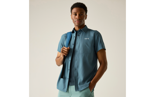 Mann in blauem Kurzarmhemd über T‑Shirt, hält einen Rucksackriemen über der Schulter, eine Hand in der Hosentasche, heller einfarbiger Hintergrund