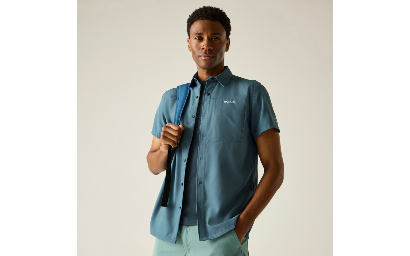 Mann in blauem Kurzarmhemd über T‑Shirt, hält einen Rucksackriemen über der Schulter, eine Hand in der Hosentasche, heller einfarbiger Hintergrund