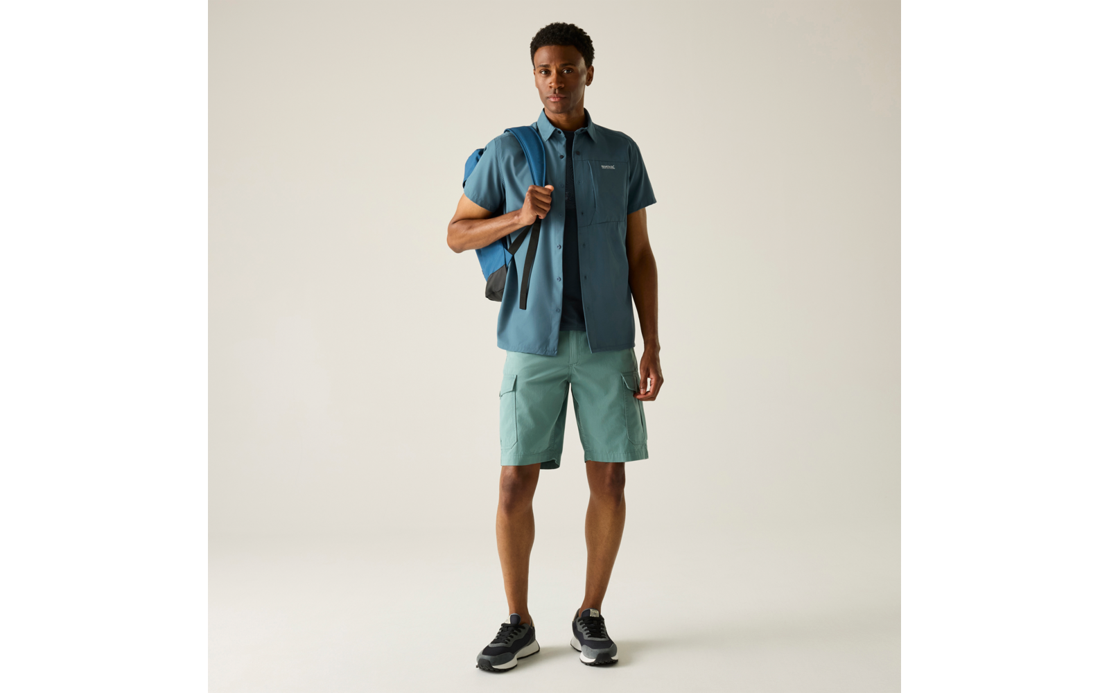 Vollkörperaufnahme einer stehenden Person, frontal, blaues kurzärmeliges Hemd, türkisfarbene Cargo-Shorts, Rucksack über der rechten Schulter, schwarze Sneaker, heller neutraler Hintergrund