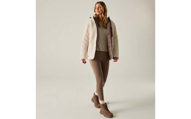 Persona con capelli biondi lunghi in piedi, indossa una giacca in pile beige chiaro con fodera rosa, una maglia beige, leggings marroni e stivaletti marroni su sfondo chiaro uniforme