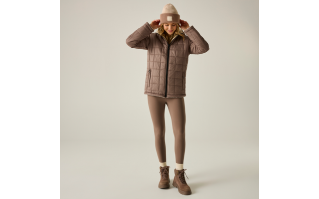 Persona in piedi davanti a uno sfondo chiaro uniforme, indossa un berretto di maglia beige, una giacca trapuntata marrone, leggings beige e stivaletti marroni con lacci, entrambe le mani sul berretto