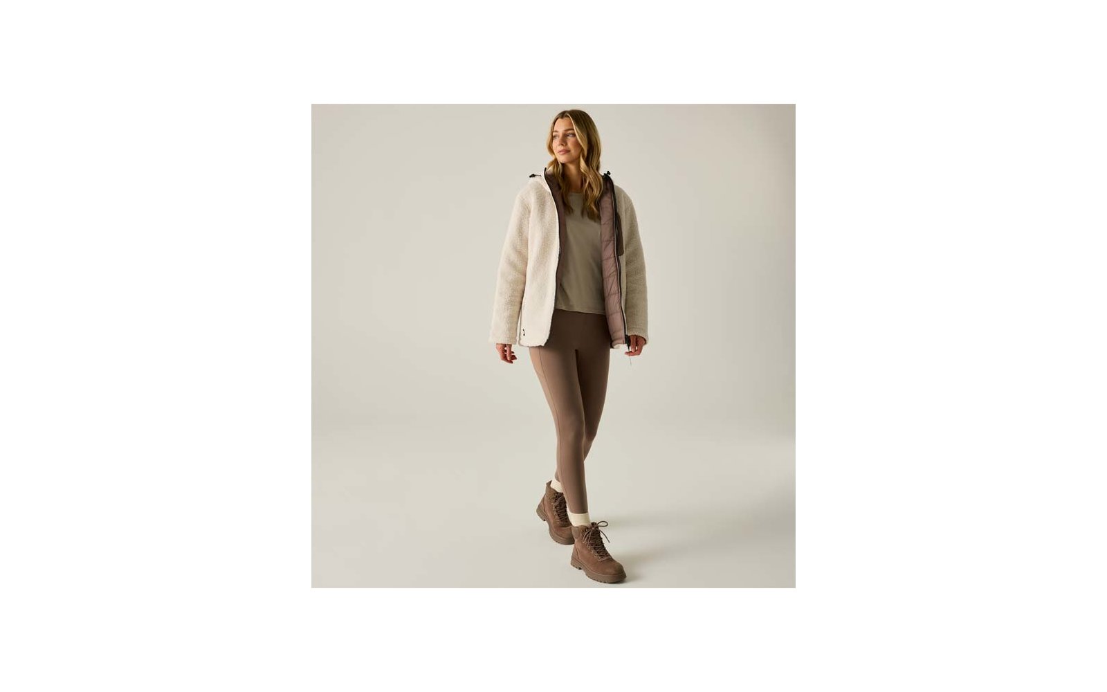 Persona con capelli biondi lunghi in piedi, indossa una giacca in pile beige chiaro con fodera rosa, una maglia beige, leggings marroni e stivaletti marroni su sfondo chiaro uniforme