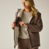 Persona apre giacca trapuntata marrone con fodera chiara in shearling, indossa maglia crema e leggings marroni, guarda verso il basso