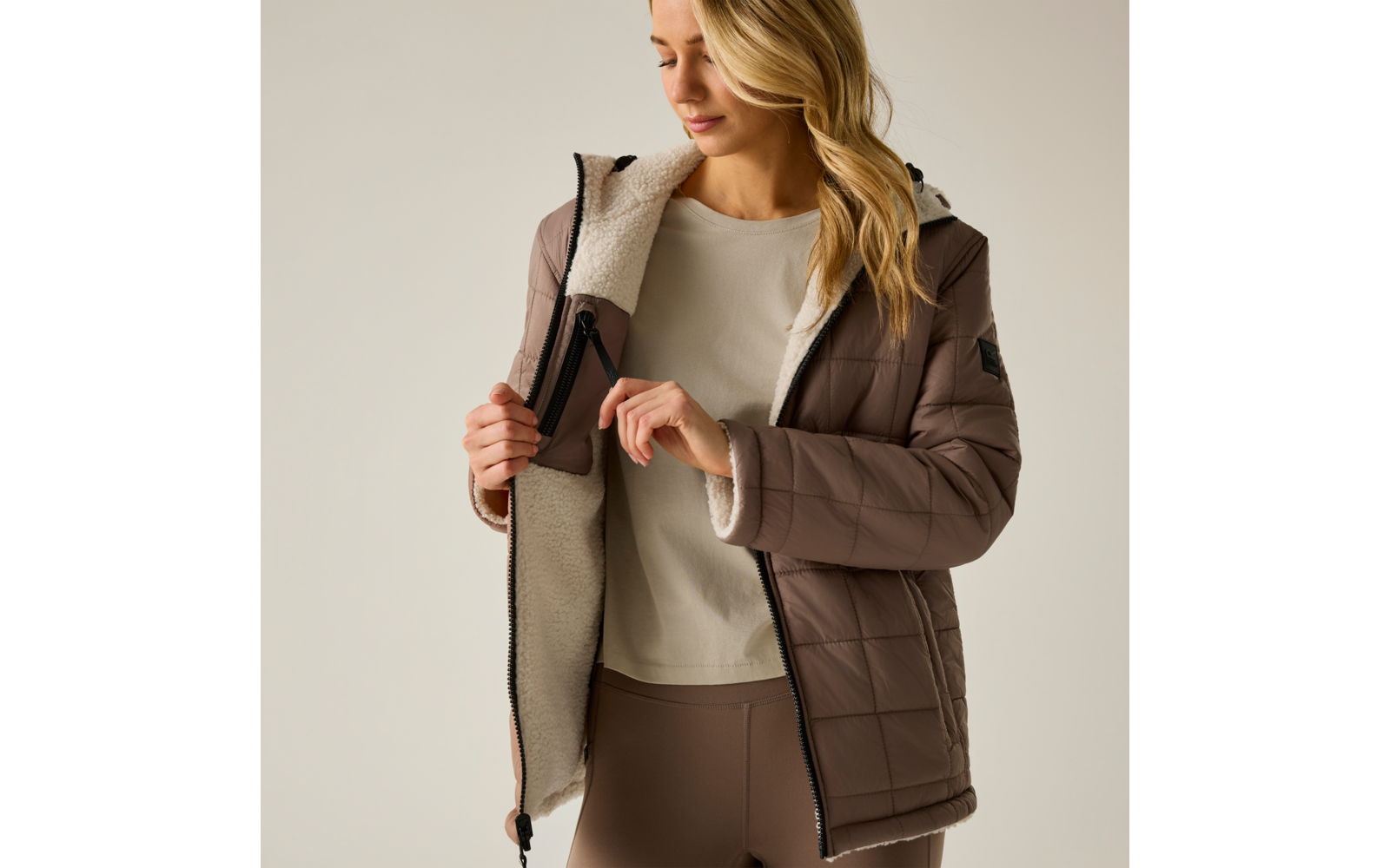 Persona apre giacca trapuntata marrone con fodera chiara in shearling, indossa maglia crema e leggings marroni, guarda verso il basso
