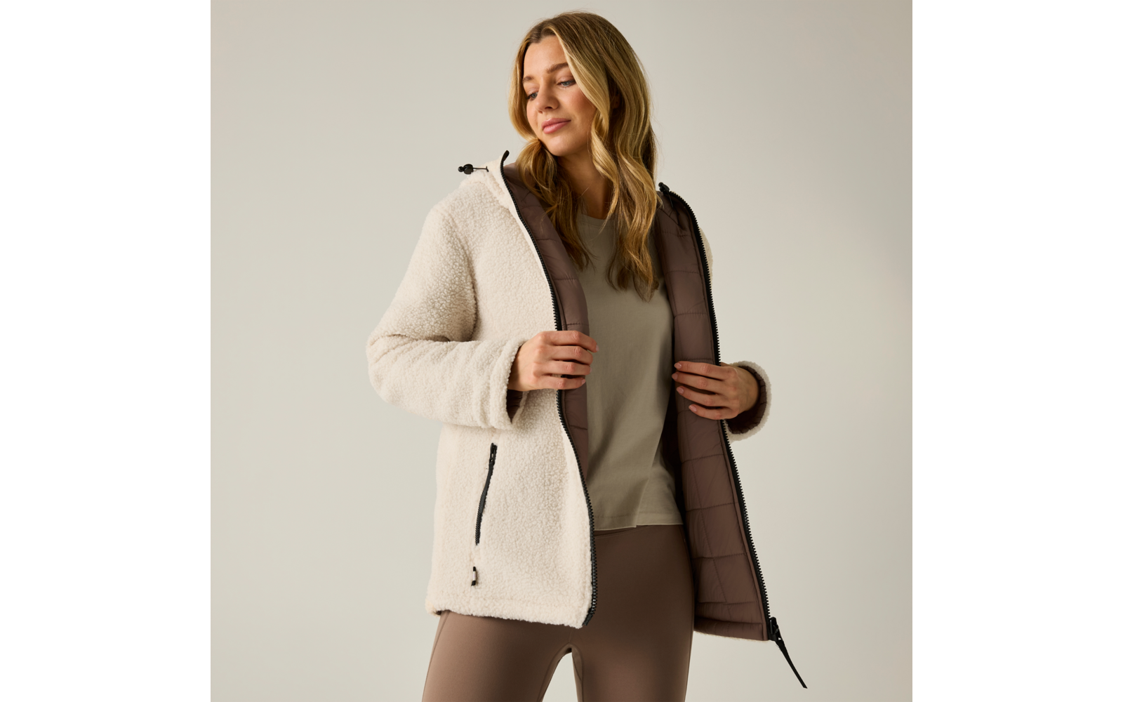 Donna con giacca sherpa crema con cappuccio, giacca aperta che mostra fodera trapuntata marrone, indossa maglia beige e leggings marroni, in piedi davanti a sfondo neutro e tiene i bordi della giacca