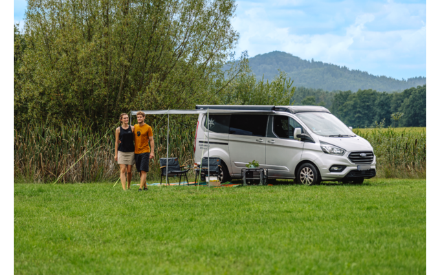 Zwei Personen stehen nebeneinander vor einem geparkten Wohnmobil auf einer Wiese; Markise ausgefahren, zwei Campingstühle und ein kleiner Tisch neben dem Fahrzeug, Bäume und Hügel im Hintergrund, bewölkter Himmel