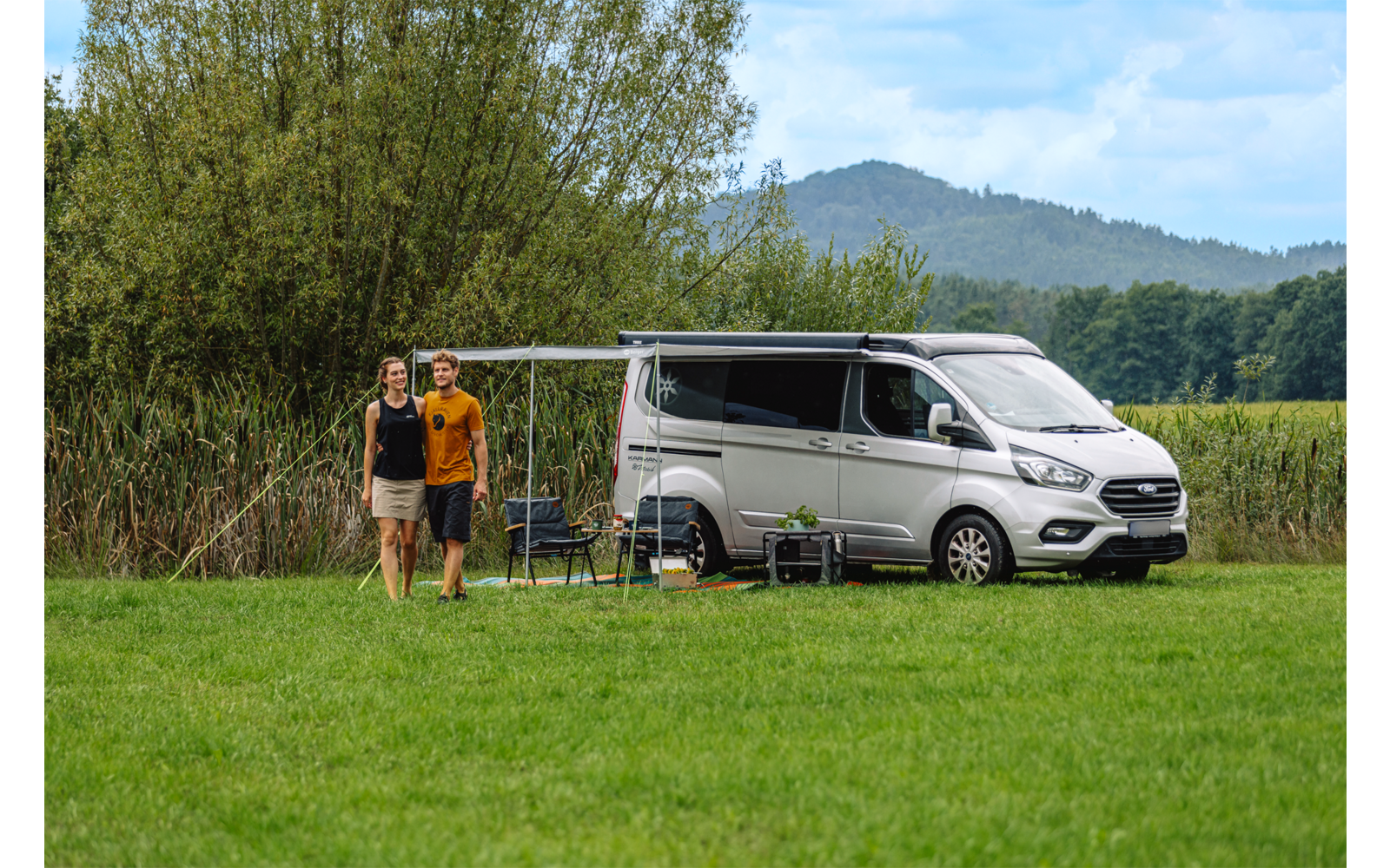 Zwei Personen stehen nebeneinander vor einem geparkten Wohnmobil auf einer Wiese; Markise ausgefahren, zwei Campingstühle und ein kleiner Tisch neben dem Fahrzeug, Bäume und Hügel im Hintergrund, bewölkter Himmel