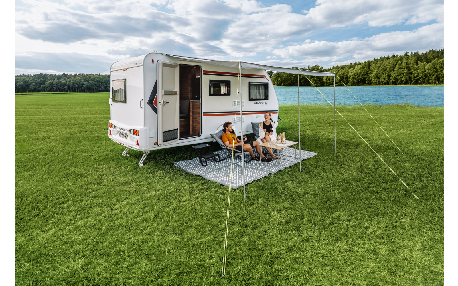 Wohnwagen auf grüner Wiese neben einem See; vor dem geöffneten Eingang sitzen zwei Personen auf Campingstühlen unter einer ausgefahrenen Markise auf einer karierten Decke, neben ihnen ein kleiner Tisch mit Bechern; im Hintergrund Wald und bewölkter H