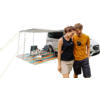 Weißer Van mit ausgeklapptem Sonnensegel; darunter gestreifte bunte Bodenmatte, zwei Campingstühle, kleiner Tisch mit Ausrüstung; rechts gehen ein Mann in gelbem T‑Shirt und eine Frau in schwarzem Top arm in arm