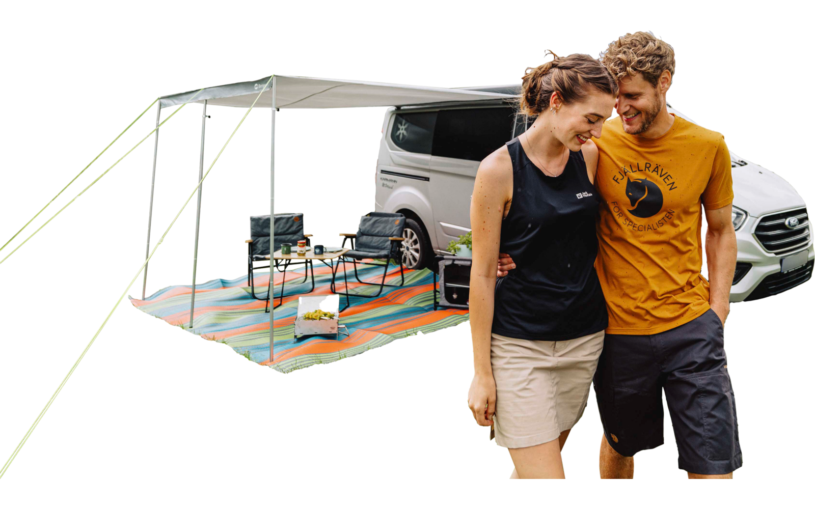 Weißer Van mit ausgeklapptem Sonnensegel; darunter gestreifte bunte Bodenmatte, zwei Campingstühle, kleiner Tisch mit Ausrüstung; rechts gehen ein Mann in gelbem T‑Shirt und eine Frau in schwarzem Top arm in arm