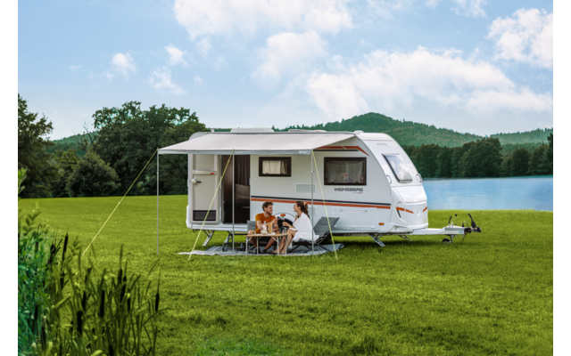 Weißer Wohnwagen mit ausgefahrenem Sonnendach auf grüner Wiese am See, drei Personen sitzen an einem Campingtisch unter dem Dach