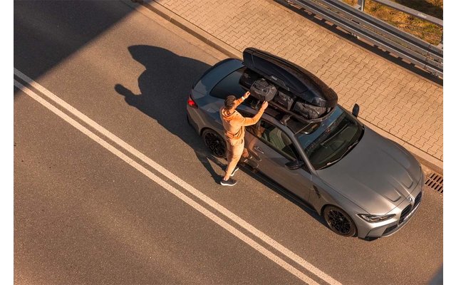 Vogelperspektive auf ein graues Auto am Straßenrand; eine Person steht daneben und greift in einen geöffneten Dachkoffer; weiße Fahrbahnmarkierungen und Schatten sichtbar