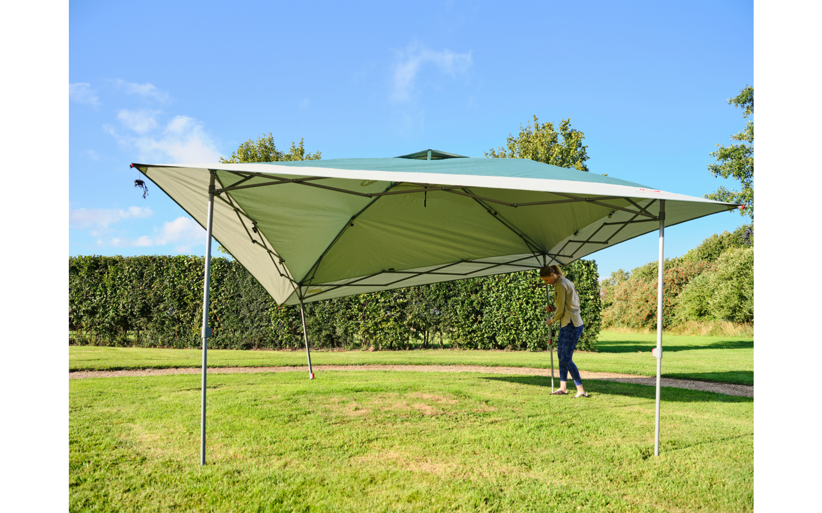 Grünes Faltzelt mit vier Metallstützen auf Rasen, Person stellt eine Stange ein, Hecke und Bäume im Hintergrund, blauer Himmel