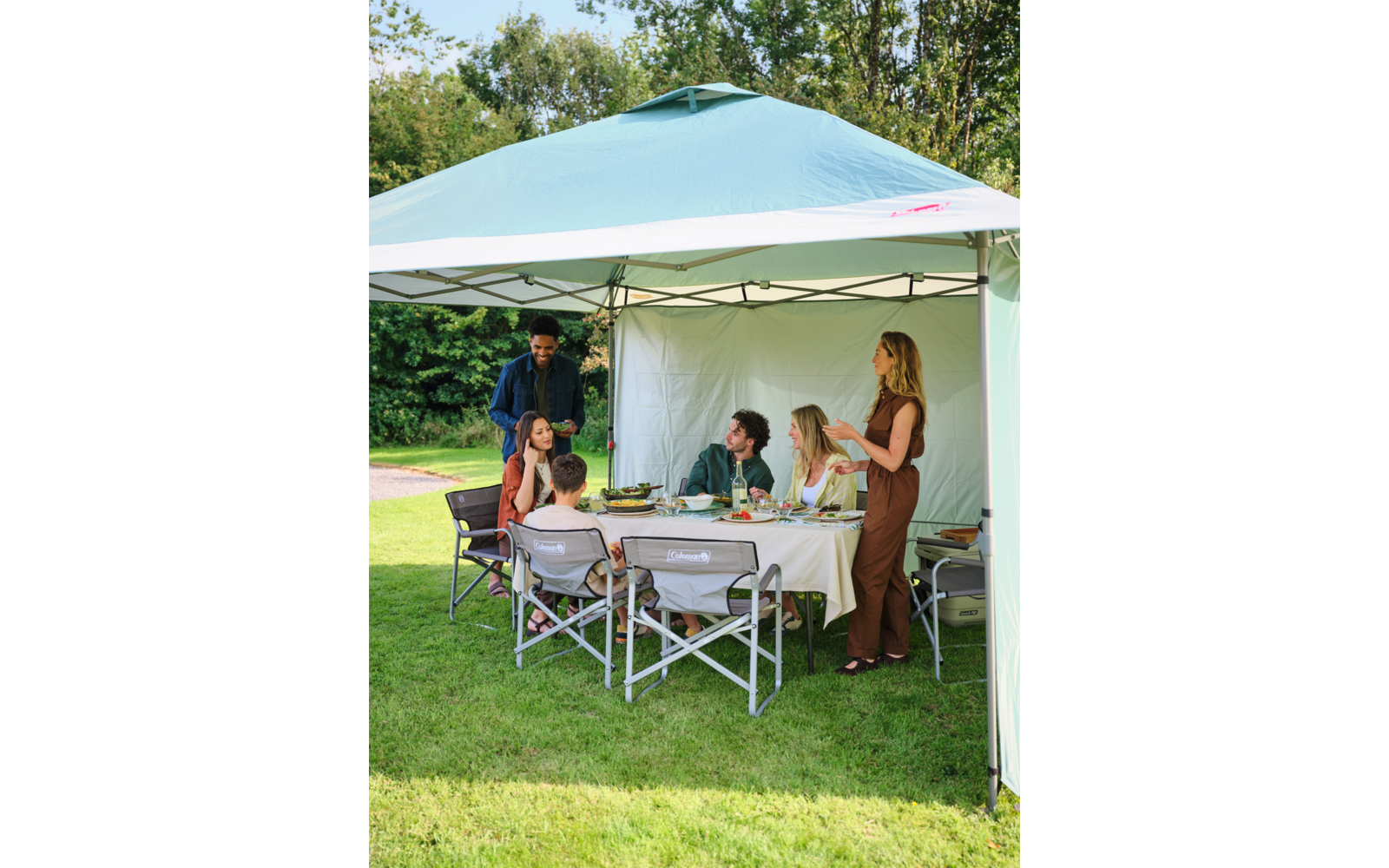 Sechs Personen sitzen und stehen unter einem hellblauen Pavillon im Garten an einem gedeckten Tisch mit Tellern und Getränken