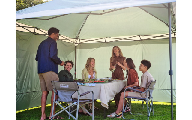 Sechs Personen unter einem grünen Pavillon: fünf sitzen an einem Tisch mit weißer Tischdecke, einer steht. Auf dem Tisch Teller, Gläser, Schalen und eine Flasche; Klappstühle auf Rasen