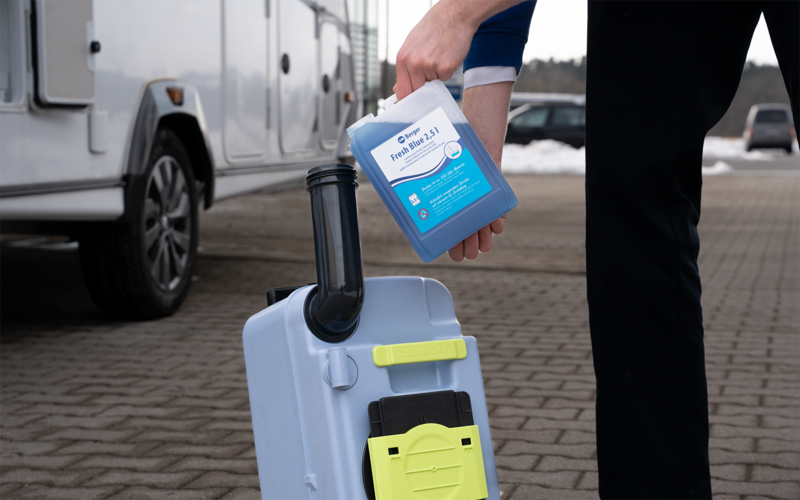Person gießt blauen Kanister in weißen Kunststoffbehälter mit schwarzem Rohr und gelbem Verschluss; Wohnmobil und gepflasterter Boden im Hintergrund
