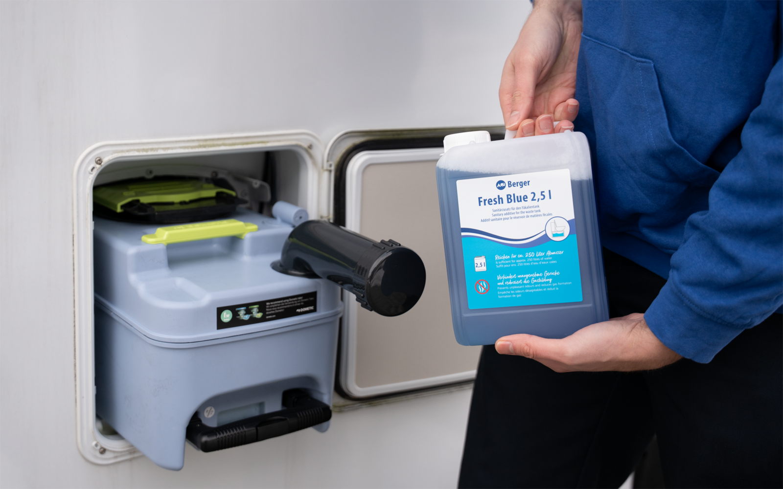 Hand hält blauen Kunststoffkanister mit Etikett 'Fresh Blue 2,5 l' neben offenem weißem Schrankfach mit grauem Behälter, schwarzem Griff und grüner Klappe