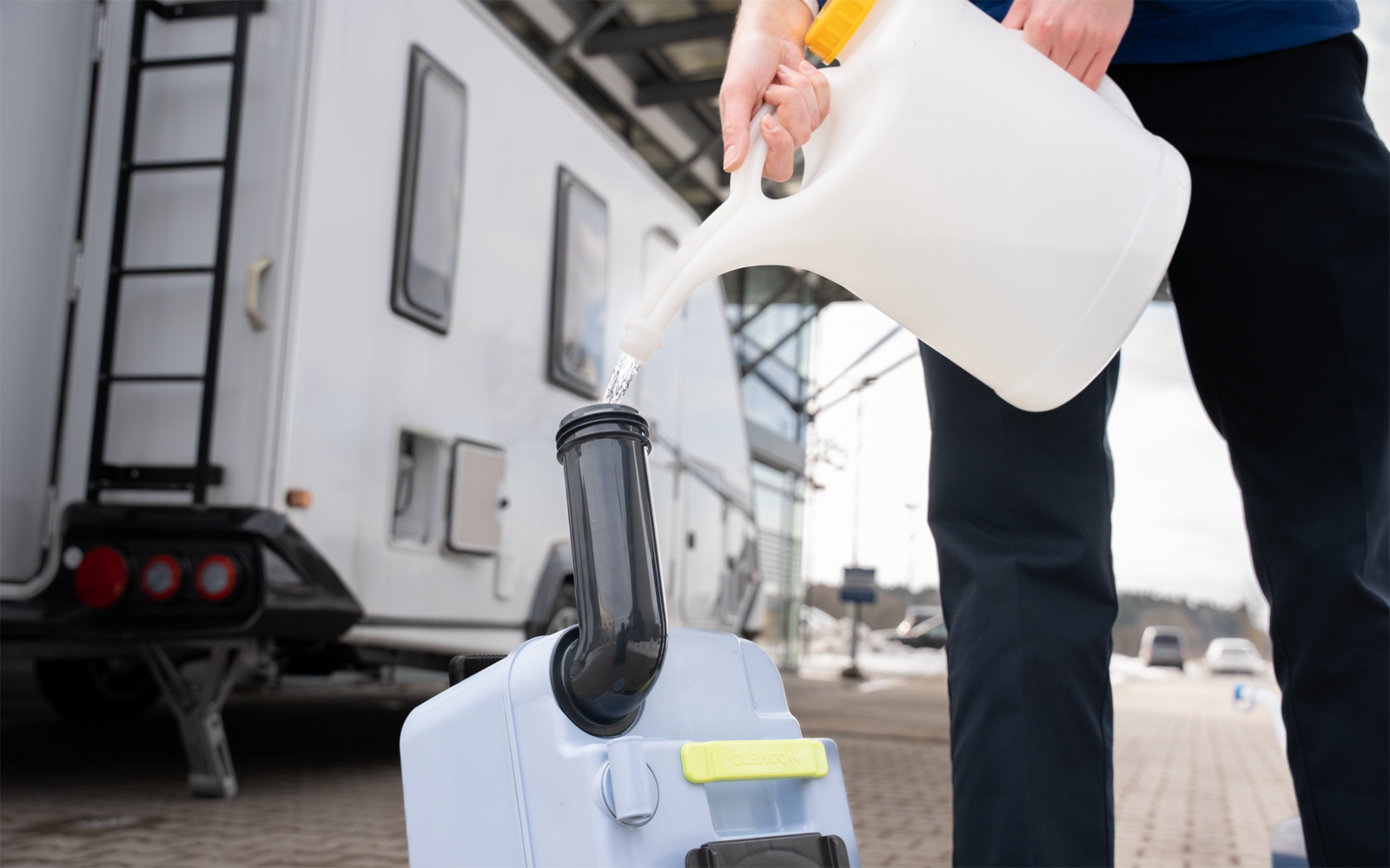 Person gießt Flüssigkeit aus einem weißen Kunststoffkanister in einen grauen Trichter an einem Kunststoffbehälter; im Hintergrund ein Wohnwagen auf gepflastertem Boden