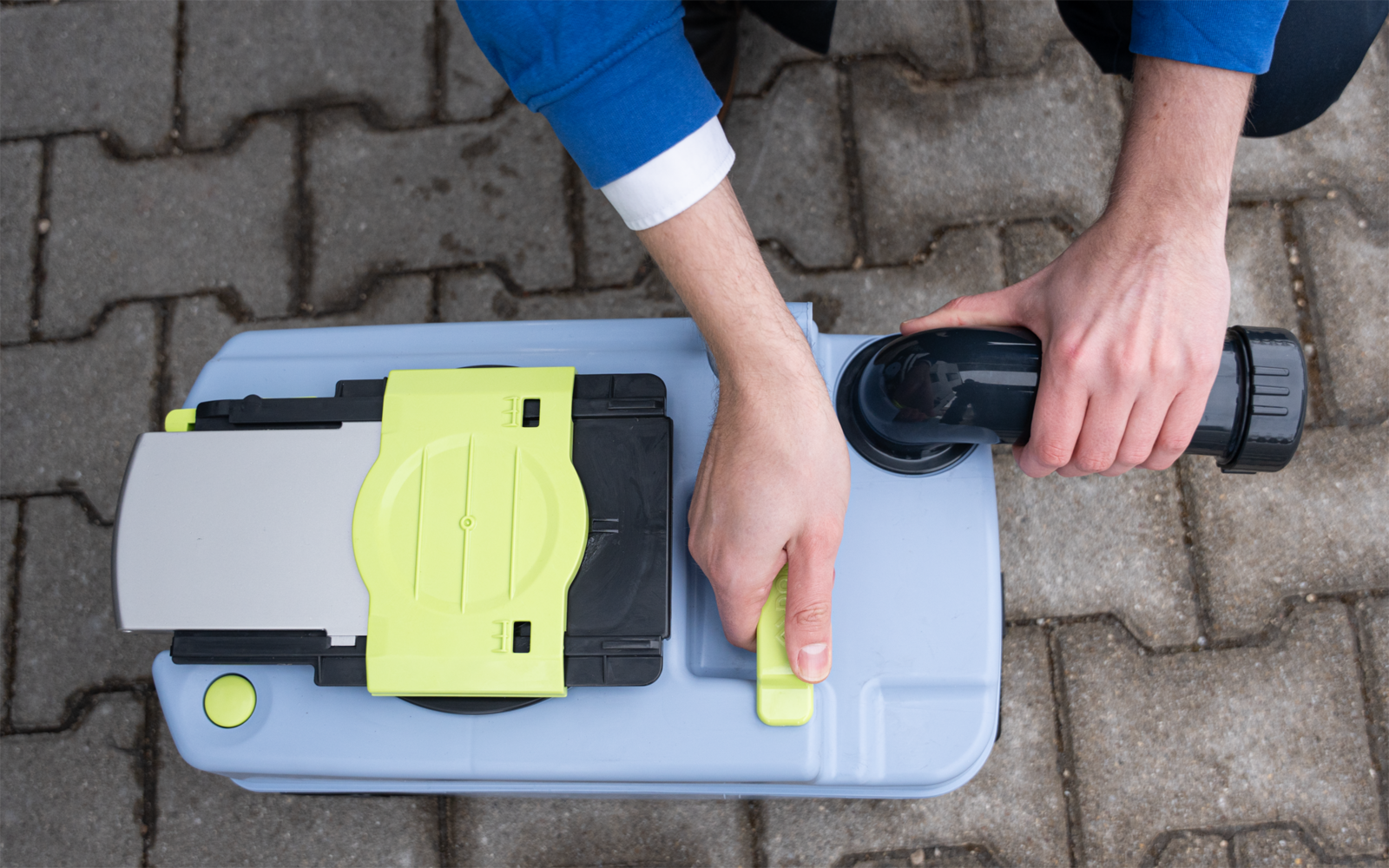 Draufsicht: zwei Hände in blauen Ärmeln halten einen schwarzen Schlauch und betätigen einen limettengrünen Verschluss an einem hellgrauen Kunststoffgerät auf Pflasterboden