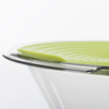 Bol en verre transparent avec couvercle en silicone vert à rainures concentriques et une cuillère métallique sur le rebord, fond blanc