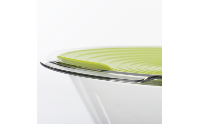 Bol en verre transparent avec couvercle en silicone vert à rainures concentriques et une cuillère métallique sur le rebord, fond blanc