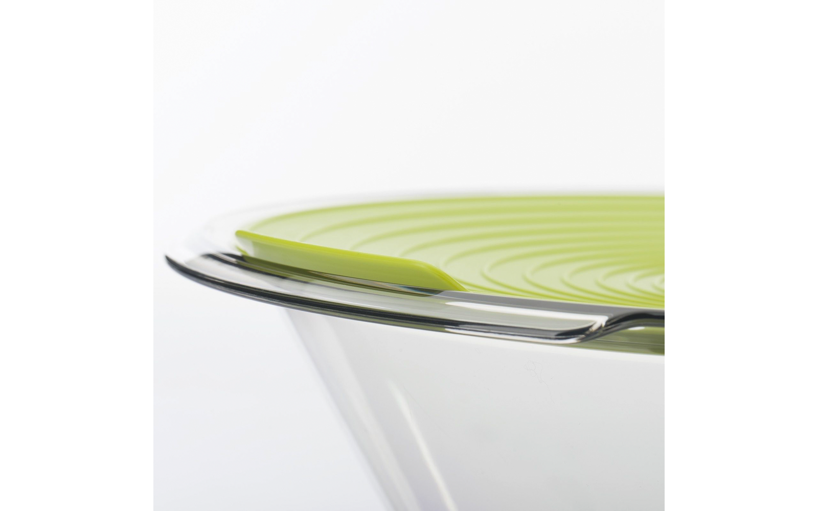 Bol en verre transparent avec couvercle en silicone vert à rainures concentriques et une cuillère métallique sur le rebord, fond blanc