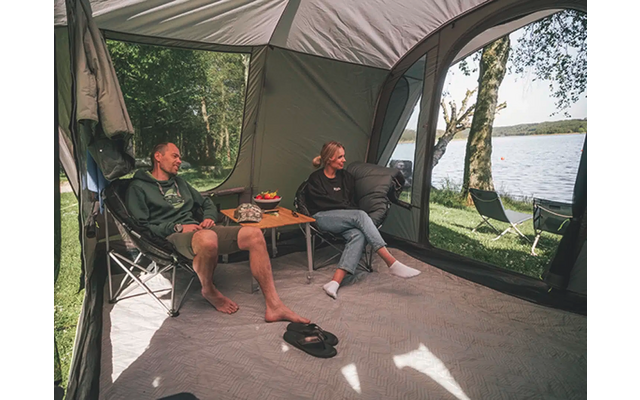 Zwei Männer sitzen auf Campingstühlen unter einem Zelt neben einem Wohnmobil, mit Blick auf einen See und Bäume