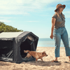 Frau mit Sonnenhut, Sonnenbrille, T-Shirt und Jeans steht barfuß auf Sand; Hund läuft aus einer kleinen dunklen Hundeterrasse/ Hundezelt; Meer und Himmel im Hintergrund