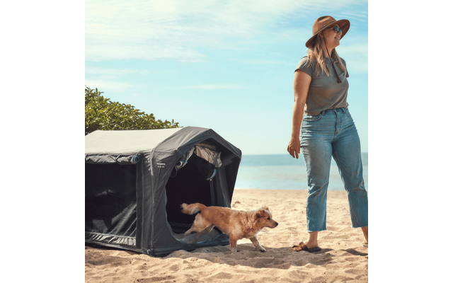 Frau mit Sonnenhut, Sonnenbrille, T-Shirt und Jeans steht barfuß auf Sand; Hund läuft aus einer kleinen dunklen Hundeterrasse/ Hundezelt; Meer und Himmel im Hintergrund