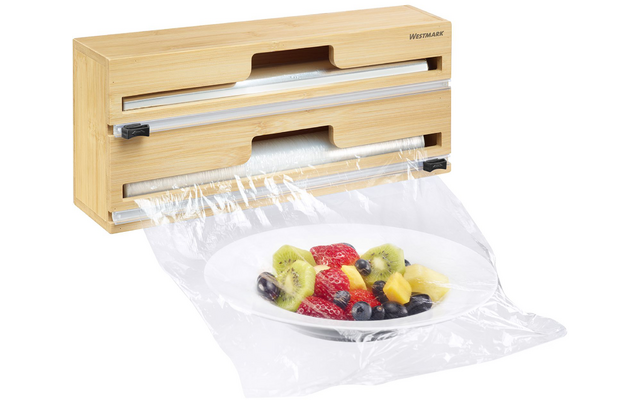 Dispensador de madera clara con dos rollos, film plástico transparente cubre un plato blanco con fresas, rodajas de kiwi, trozos de piña y arándanos