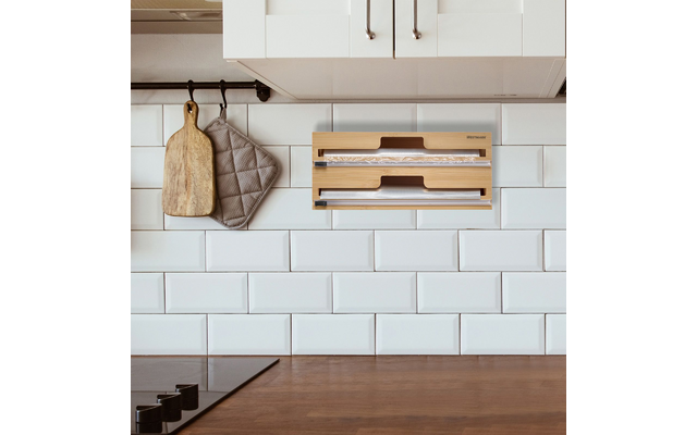 Revestimiento de azulejos blancos tipo metro, encimera de madera, placa de cocción negra a la izquierda con mandos redondos, soporte mural de madera con dos rollos visibles, tabla de cortar de madera y agarradera gris colgadas en una barra, armarios altos blancos con tiradores metálicos
