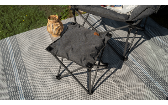 Ein grauer Klapphocker auf einer gestreiften grauen Decke neben einem geflochtenen Korb und einem Klappstuhl