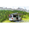 Campingbus mit ausgefahrener Markise und zwei Personen, eine sitzt auf einem Klappstuhl, in einer grünen Wiese vor bewaldeten Bergen