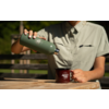 Buste d'une personne en chemise vert clair versant d'une thermos verte dans une tasse rouge sur une table en bois ; mains avec vernis blanc visibles ; banc et végétation flous en arrière-plan
