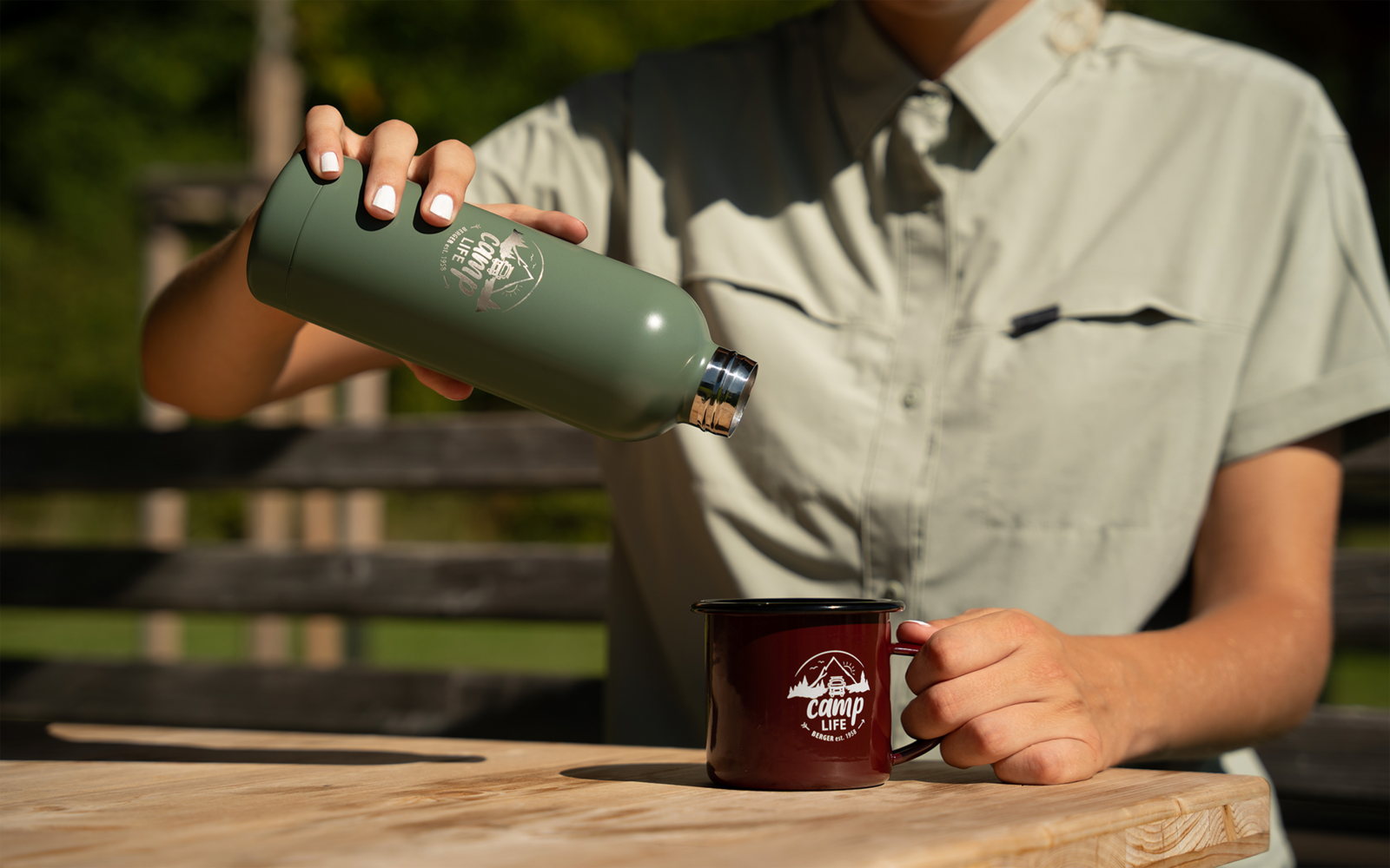 Buste d'une personne en chemise vert clair versant d'une thermos verte dans une tasse rouge sur une table en bois ; mains avec vernis blanc visibles ; banc et végétation flous en arrière-plan