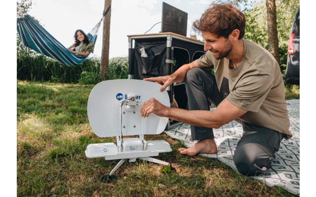 Un homme assis par terre utilise une machine à coudre en plein air, avec une personne allongée dans un hamac en arrière-plan