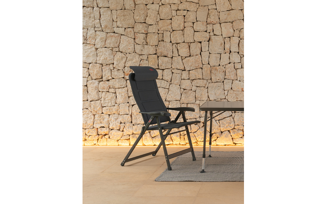 Schwarzer klappbarer Liegesessel mit Kopfstütze und Armlehnen neben einem Tisch auf einem hellen Teppich vor einer beigen Natursteinwand mit warmem Bodenlicht
