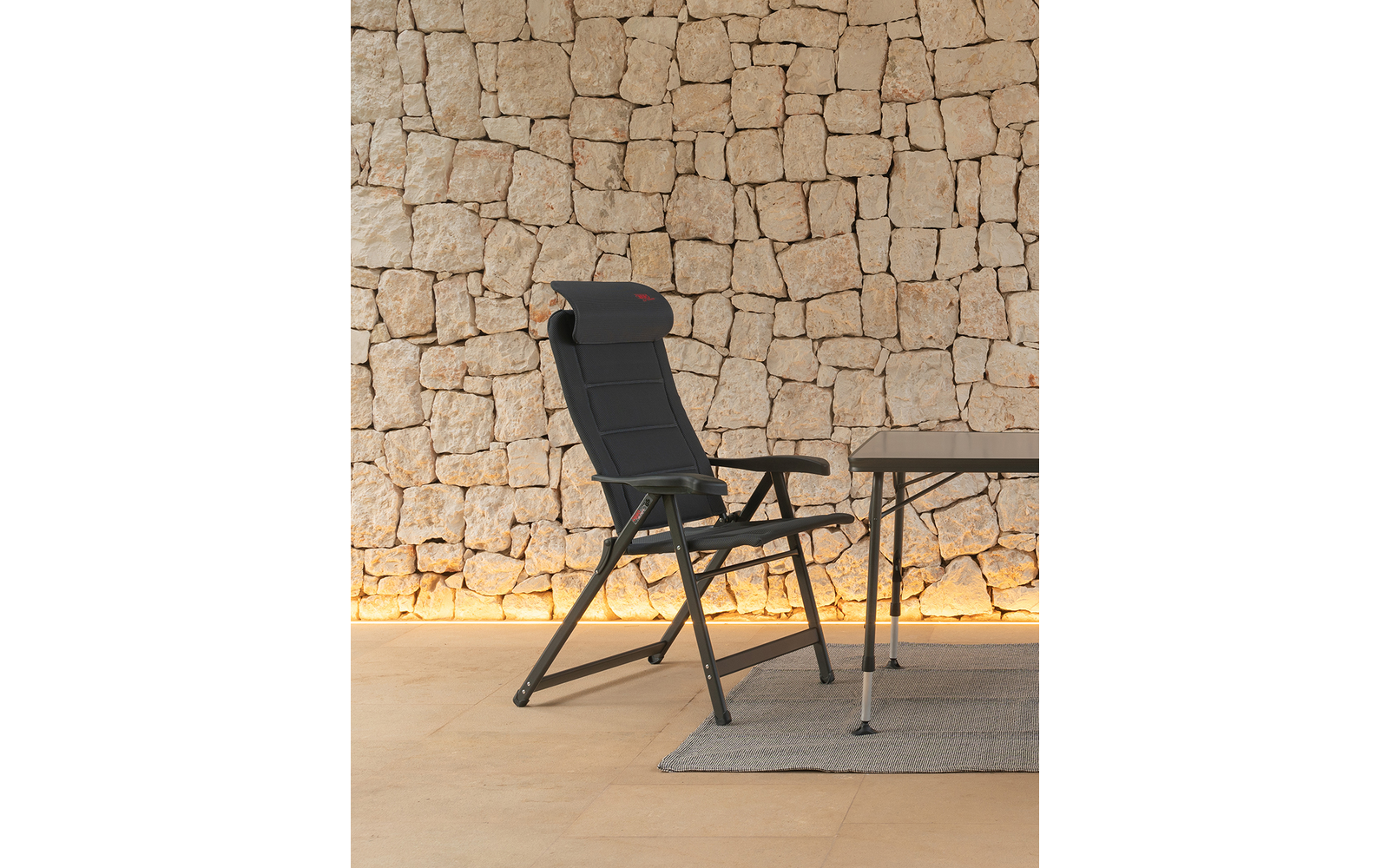Schwarzer klappbarer Liegesessel mit Kopfstütze und Armlehnen neben einem Tisch auf einem hellen Teppich vor einer beigen Natursteinwand mit warmem Bodenlicht
